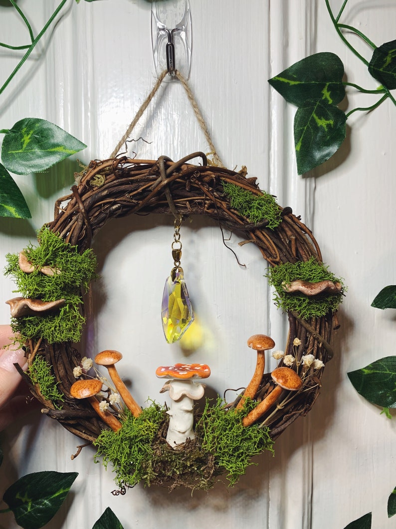 May include: A brown twig wreath with green moss, small white flowers, and brown mushrooms. A yellow crystal hangs from the wreath.