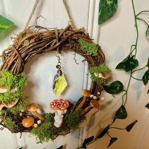 May include: A brown twig wreath decorated with moss, small white flowers, and miniature mushrooms. A crystal prism hangs from the wreath.