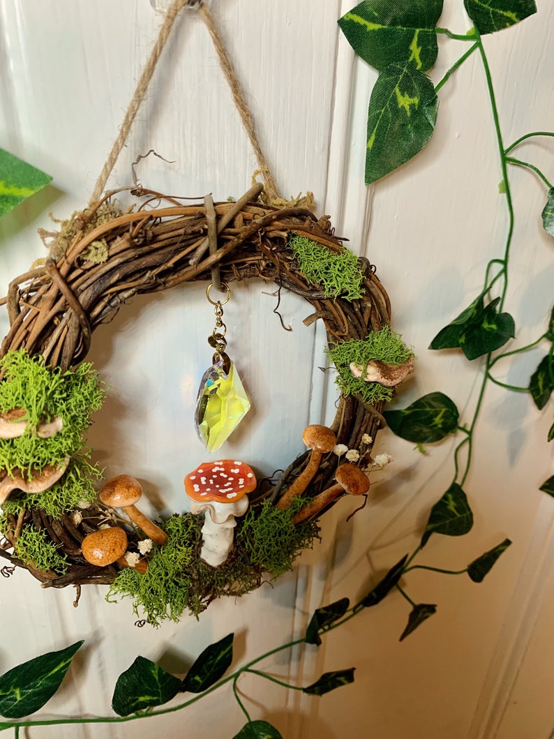 May include: A brown twig wreath decorated with moss, small white flowers, and miniature mushrooms. A crystal prism hangs from the wreath.