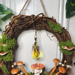 May include: A brown twig wreath with moss, small mushrooms, and a crystal pendant hanging in the center. The wreath is decorated with white flowers and has a rustic, woodland theme.