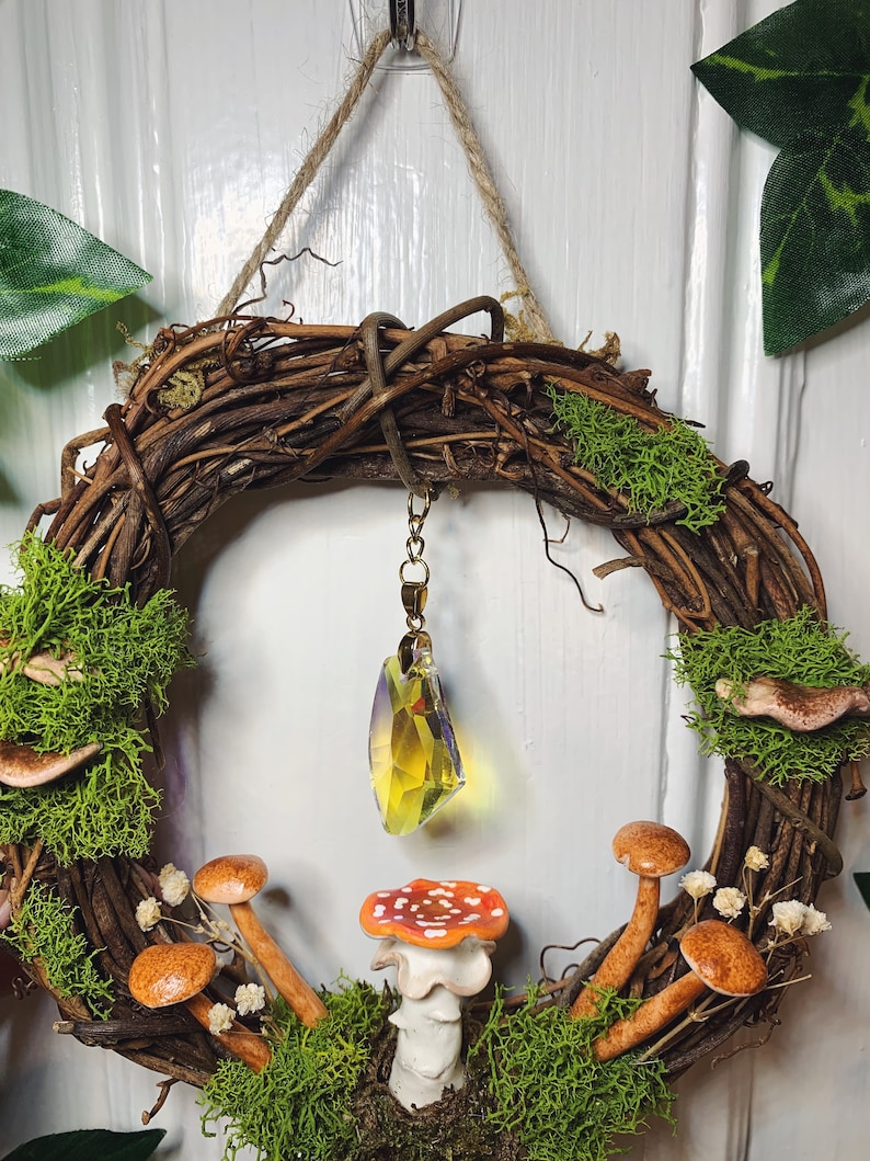 May include: A brown twig wreath with moss, small mushrooms, and a crystal pendant hanging in the center. The wreath is decorated with white flowers and has a rustic, woodland theme.