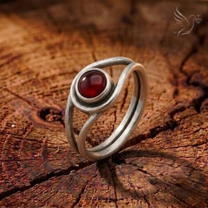 May include: A silver ring featuring a smooth, round, deep red stone set in a bezel. The band is constructed from two separate silver wires that curve and meet to hold the central stone, creating an open, layered appearance with a metallic finish.