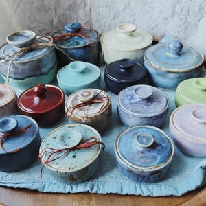 May include: A collection of small, round ceramic jars with matching lids in various colors including blue, red, green, purple, and cream. Some jars feature speckled glazes and are tied with thin twine or string, arranged together on a light blue cloth.