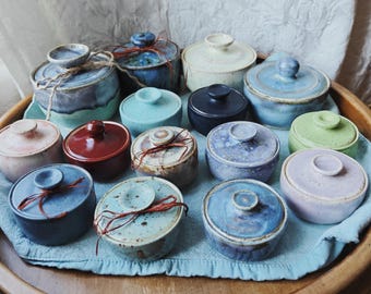 May include: A collection of small, round ceramic jars with matching lids in various colors including blue, red, green, purple, and cream. Some jars feature speckled glazes and are tied with thin twine or string, arranged together on a light blue cloth.