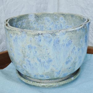 May include: A round ceramic planter with a scalloped rim and a matching saucer. The exterior features a speckled glaze with mottled patches of light blue and cream, creating a textured appearance. The base of the planter is a solid, unglazed clay.