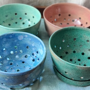 May include: Four ceramic bowls in shades of blue, green, and pink. Each bowl has a series of small holes. The bowls are arranged on a light-colored surface, suggesting a kitchen or dining setting. The bowls are likely for food preparation or serving.