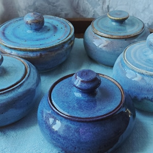 May include: A collection of five round ceramic lidded jars featuring a glossy glaze in various shades of blue, ranging from deep indigo to light sky blue. Each jar has a matching lid with a small knob handle and a smooth, rounded silhouette.
