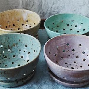 May include: Four ceramic berry bowls in yellow, mint green, speckled blue-green, and light purple. Each bowl features a rounded shape with multiple drainage holes distributed across the base and sides, and includes a matching ceramic saucer underneath.