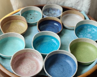 May include: A collection of eleven small, round ceramic bowls with speckled, off-white exteriors and various interior glaze colors including shades of blue, green, pink, yellow, and white, arranged together on a circular wooden tray.