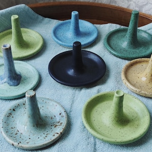 May include: A collection of eight ceramic ring holders, each featuring a circular base and a central vertical post. The holders come in various speckled finishes, including light green, blue, dark green, black, yellow, and pale blue with brown spots.