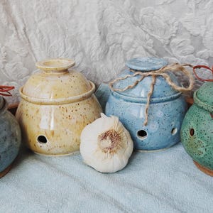 May include: Four round ceramic garlic keepers with lids, each featuring small ventilation holes near the base. The containers are glazed in speckled blue, yellow, light blue, and green, with thin brown twine tied around the lids of the latter two.