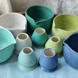 May include: A collection of handmade ceramic bowls and bud vases in various shades of blue, green, and cream. The pottery has a speckled texture. Some bowls have a spout. The vases are small and rounded.