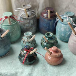 May include: A collection of ceramic honey pots in various sizes and colors, including blue, green, pink, and grey. Each pot features a matching lid with a hole for a wooden honey dipper. Some pots are tied with thin ribbons in red or white.