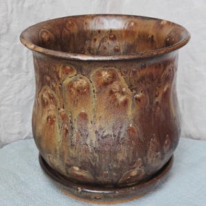 May include: A round ceramic planter with a wide, flared rim and a matching circular saucer. The exterior features a mottled glaze with blended tones of deep chocolate brown, warm tan, and golden yellow, creating a textured, drip-like appearance.
