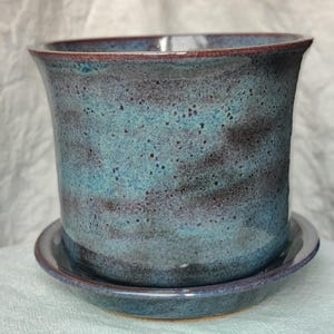 May include: A handcrafted ceramic planter with a matching saucer. The planter is cylindrical with a flared rim, and the exterior features a mottled blue and dark brown glaze. The rim has a reddish-brown accent. The saucer is round and glazed to match.