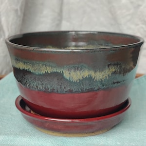 May include: A handcrafted ceramic planter with a matching saucer. The planter features a rich, glossy glaze in shades of burgundy, brown, teal, and yellow. The bowl-shaped planter sits atop a round saucer, perfect for indoor plants.