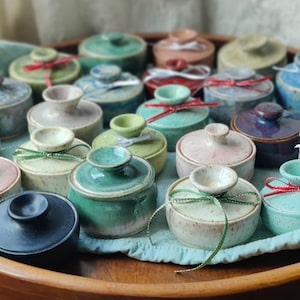 May include: A collection of small, ceramic lidded jars in various pastel colors, including pink, blue, and green. Each jar is adorned with a small ribbon, and they are arranged on a wooden tray with a blue cloth.