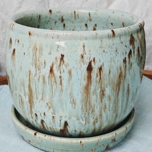 May include: A round ceramic planter with a light mint green glaze featuring vertical brown drip patterns. The planter has a matching circular saucer base with a raised rim, creating a cohesive set for holding small plants or decorative items.