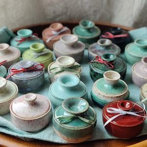 May include: A collection of small, ceramic lidded jars in various colors, including teal, pink, and green, arranged on a wooden tray. Each jar is adorned with a decorative ribbon. The jars have a speckled finish.