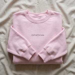 May include: A light pink sweatshirt with the word "ESTHETICIAN" printed in a small, dark font across the chest. The sweatshirt is folded neatly, with ribbed cuffs and a crew neck. The fabric appears soft and comfortable.