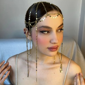 Może przedstawiać: Złota główka z łańcucha z wiszącymi łańcuchami z małych złotych kółek. Głowka jest noszona przez osobę o ciemnych włosach i makijażu.