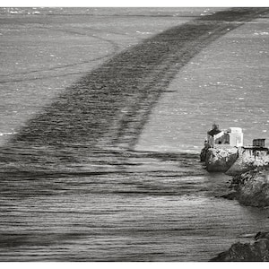 Könnte beinhalten: Ein Schwarzweißfoto eines Leuchtturms auf einer felsigen Klippe mit Blick auf das Meer. Der Leuchtturm ist ein kleines, weißes Gebäude mit einem roten Dach. Das Meer ist ruhig und der Himmel ist bewölkt.