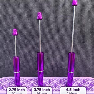 Könnte beinhalten: Drei violette Stiftverlängerungen zum Auffädeln in verschiedenen Längen: 70 mm, 95 mm und 114 mm. Jede Verlängerung hat einen zylindrischen Körper und eine Kappe. Der Text "Beadable Pen Extenders" steht oben.