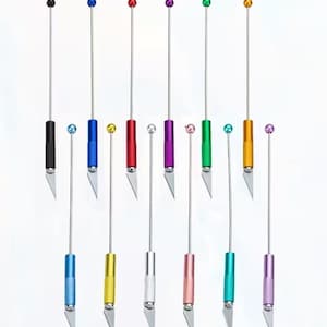 May include: A collection of craft knives with various colored handles and ball-shaped tops. The knives have a silver blade and a long, thin silver shaft. The handles are black, blue, red, purple, green, gold, silver, pink, teal, and lavender.