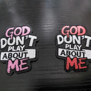 Op de afbeelding: Twee geborduurde patches met de tekst "GOD DON'T PLAY ABOUT ME". Eén patch heeft roze accenten, de andere heeft rode accenten. De patches zijn zwart met witte en gekleurde letters.