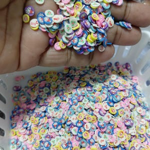 May include: A collection of colorful polymer clay slices in a white basket. The slices are round with cartoon character designs in various colors, including blue, pink, yellow, and purple. Some slices are held in a hand.