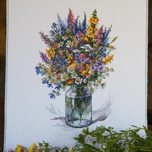 May include: A cross-stitch embroidery depicting a bouquet of wildflowers arranged in a clear glass jar. The flowers are a mix of purple, blue, yellow, and white.