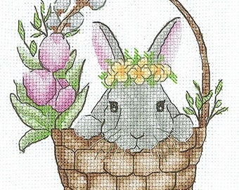 May include: A gray bunny wearing a yellow flower crown sits in a brown wicker basket with pink and green flowers.