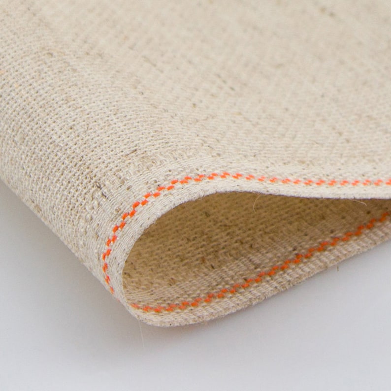 May include: Close-up of a beige fabric with an orange stitched border. The fabric has a woven texture.