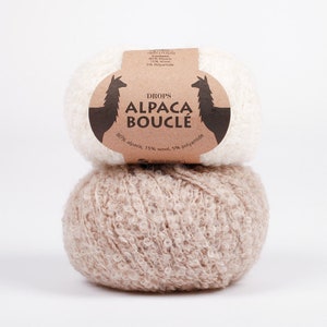 Puede incluir: Dos ovillos de lana, uno blanco y otro beige, con una etiqueta marrón que dice "DROPS ALPACA BOUCLE 80% alpaca, 15% lana, 5% poliamida".