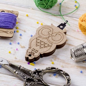 May include: Wooden owl-shaped thread holder with a metal needle threader. The holder is decorated with a simple owl design and has a hole for threading the needle. The holder is surrounded by colourful beads and yarn.