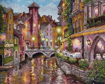 May include: A painting of a quaint European town at dusk. The scene features a canal with a bridge, colorful buildings, and streetlights. There are flowers in the foreground and a restaurant with tables and chairs on the right side of the image.