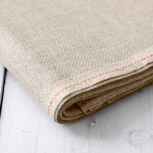 May include: A beige linen fabric with an orange stitched edge. The fabric is folded and rolled up.