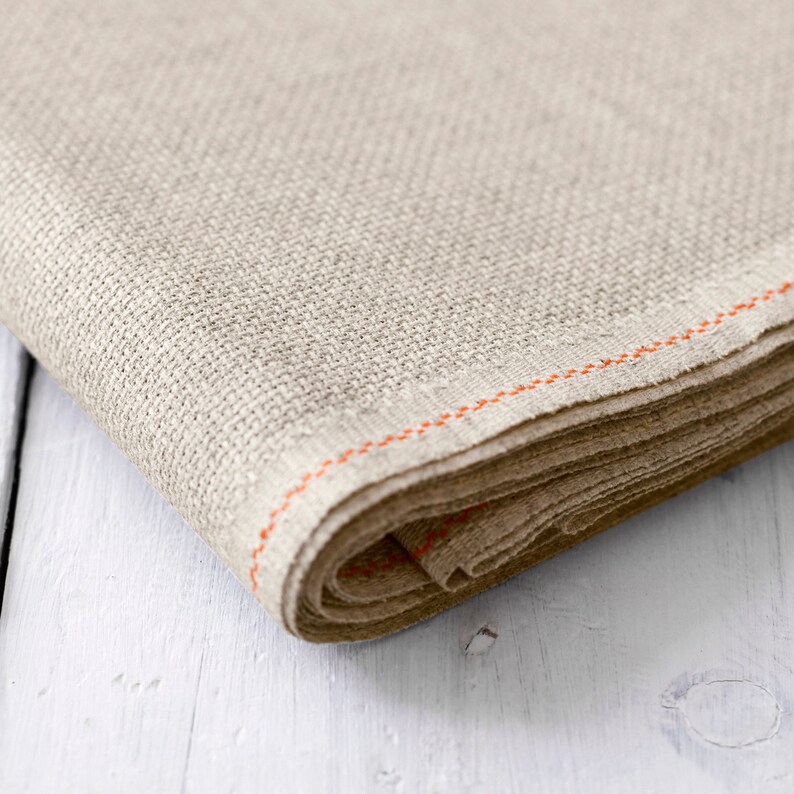 May include: A beige linen fabric with an orange stitched edge. The fabric is folded and rolled up.
