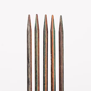 Könnte beinhalten: Fünf Stricknadeln aus Holz mit mehrfarbiger Oberfläche, darunter Brauntöne, Grüntöne und Orangetöne. Die Nadeln sind vertikal angeordnet und alle gleich lang.
