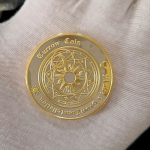 Puede incluir: Una moneda de color dorado con un centro plateado que presenta un diseño circular con un sol y una luna. La moneda tiene el texto "Tarrow Coin" y "The Love & Connection" alrededor del borde.