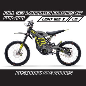 Può includere: Un kit grafico personalizzato grigio e giallo per una moto elettrica Sur-Ron Light Bee X. Il kit include un logo Fox Racing e il testo "LIGHT BEE X" e "LIE".