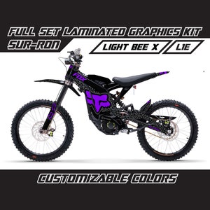 May include: A black and purple Sur-Ron Light Bee X electric dirt bike with Fox Racing graphics. The bike has a black frame, black wheels, and purple accents. The bike is shown on a white background.
