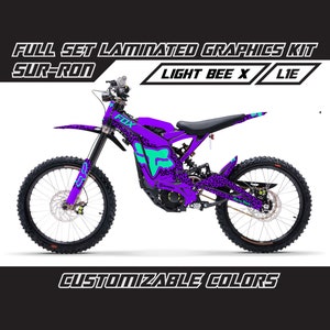 May include: A purple and green Sur-Ron Light Bee X electric dirt bike with a custom graphic kit featuring a fox logo and leopard print. The bike has black wheels and a black seat. The text "FULL SET LAMINATED GRAPHICS KIT" and "CUSTOMIZABLE COLORS" is visible in the image.