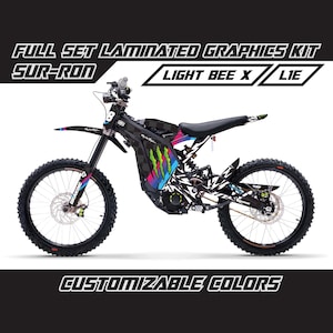 Op de afbeelding: Een zwart-witte Sur-Ron Light Bee X L1E elektrische crossmotor met een complete set gelamineerde graphics. De graphics hebben een kleurrijk ontwerp met de tekst "CUSTOMIZABLE COLORS". De motor heeft zwarte banden en een zwart zadel.