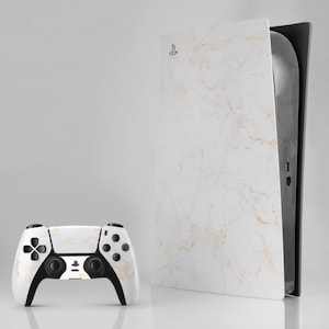 May include: A white and gold marble-effect PlayStation 5 console and matching controller. The console is upright, with the controller positioned to the left. The controller has black accents and buttons. The console has a gold PlayStation logo.
