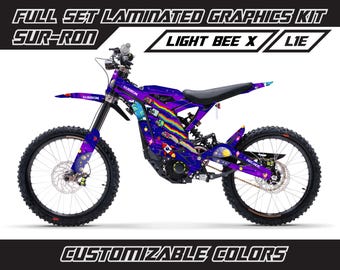 May include: A purple and blue Sur-Ron Light Bee X electric dirt bike with a space-themed graphic design. The bike has a black seat and black wheels with white spokes. The graphics include planets, stars, and a rainbow. The text "SUR-RON" and "ARCADE" are visible on the bike. The text "FULL SET LAMINATED GRAPHICS KIT" and "CUSTOMIZABLE COLORS" are visible at the top and bottom of the image.