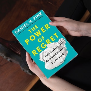 May include: A teal blue book with the title "The Power of Regret" in yellow text. The book is titled "How Looking Backward Moves Us Forward" and has a "Large Print" label. The book is being held by a person's hands.