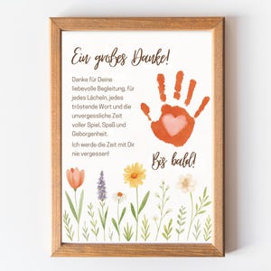 Könnte beinhalten: Ein gerahmter Druck mit einem floralen Rand und dem Text "Ein großes Danke!" in einer handschriftlichen Schriftart. Ein orangefarbener Handabdruck mit einem Herz ist abgebildet. Der Druck befindet sich in einem Holzrahmen.