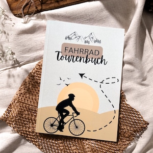 Könnte beinhalten: Ein braunes und weißes Notizbuch mit dem Text "FAHRRAD Tourenbuch" auf dem Cover. Das Cover zeigt außerdem eine Silhouette einer Person, die auf einem Weg mit einer gepunkteten Linie und einem Sonnenuntergang im Hintergrund Fahrrad fährt.