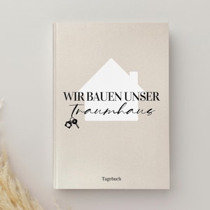 Könnte beinhalten: Ein beiges Notizbuch mit dem Text "WIR BAUEN UNSER Traumhaus" und einer weißen Hausgrafik. Das Notizbuch hat auch eine Schlüsselgrafik und das Wort "Tagebuch" unten. Das Cover hat ein mattes Finish.